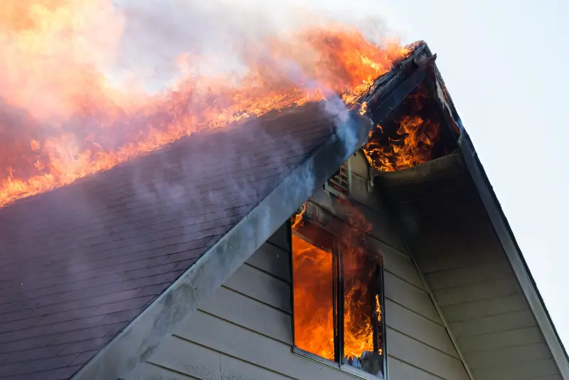 Flames and smoke engulf the roof and window of a house on fire.
