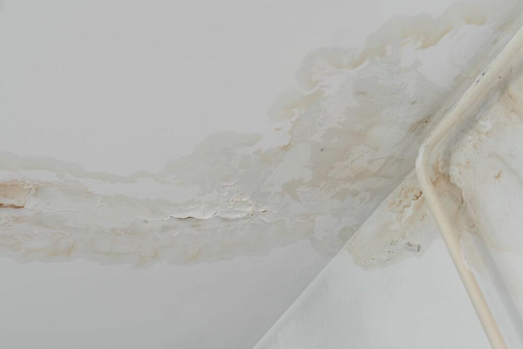 Water damage and peeling paint on a ceiling corner near a pipe.