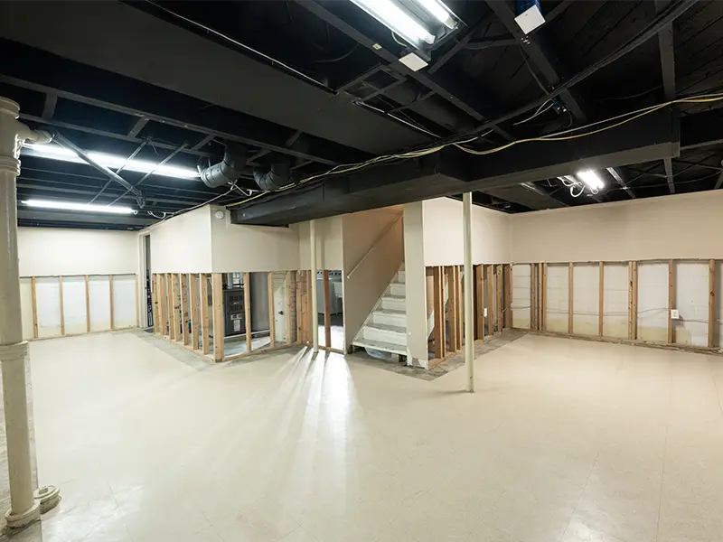 Basement with exposed wall studs, a staircase, and a black open ceiling with fluorescent lights. commercial restoration services